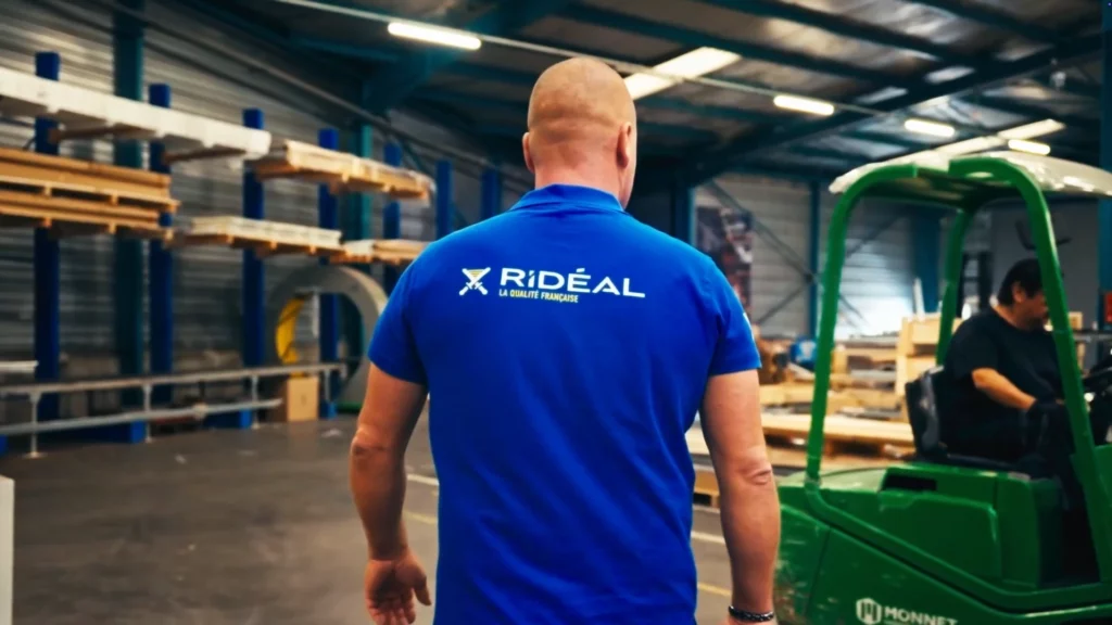 Salarie de dos en polo bleu Rideal marchant dans un atelier, chariot elevateur vert et racks de stockage